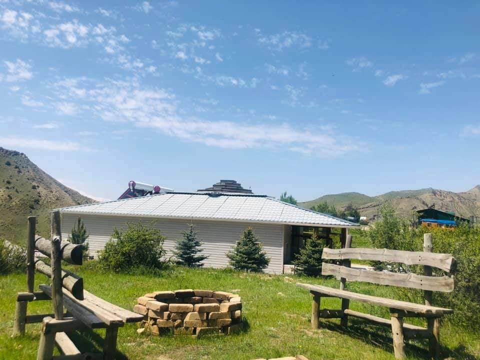 A serene outdoor area features wooden benches arranged around a stone fire pit, set against a backdrop of rolling hills and clear blue skies. Nearby, the lodge's white exterior can be seen, harmonizing with the natural landscape.
