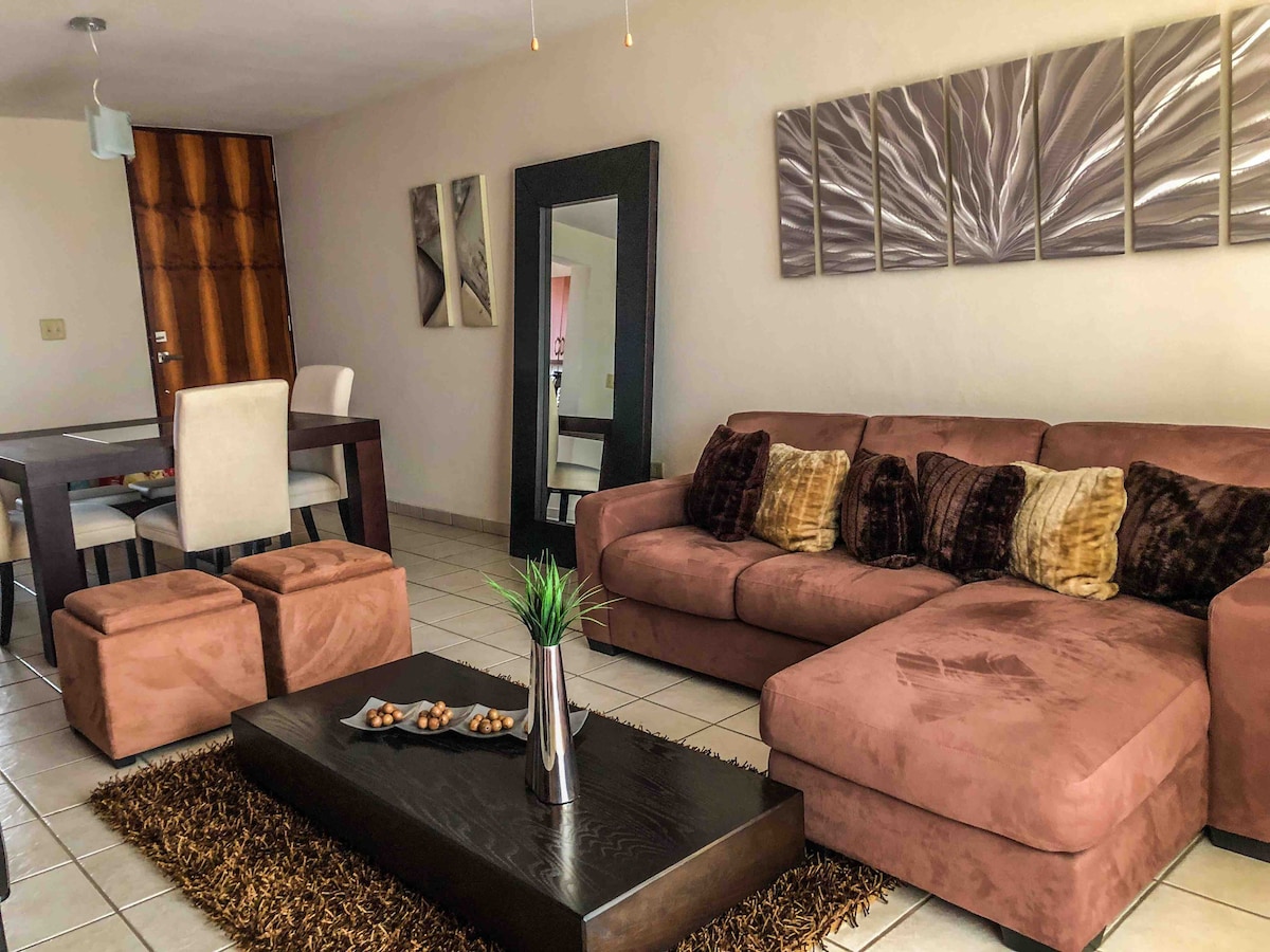 A spacious living area features a large sectional sofa adorned with decorative pillows, complemented by a sleek coffee table. A textured rug adds warmth to the tiled floor, while abstract wall art provides a modern touch. A dining table with chairs is visible in the background.