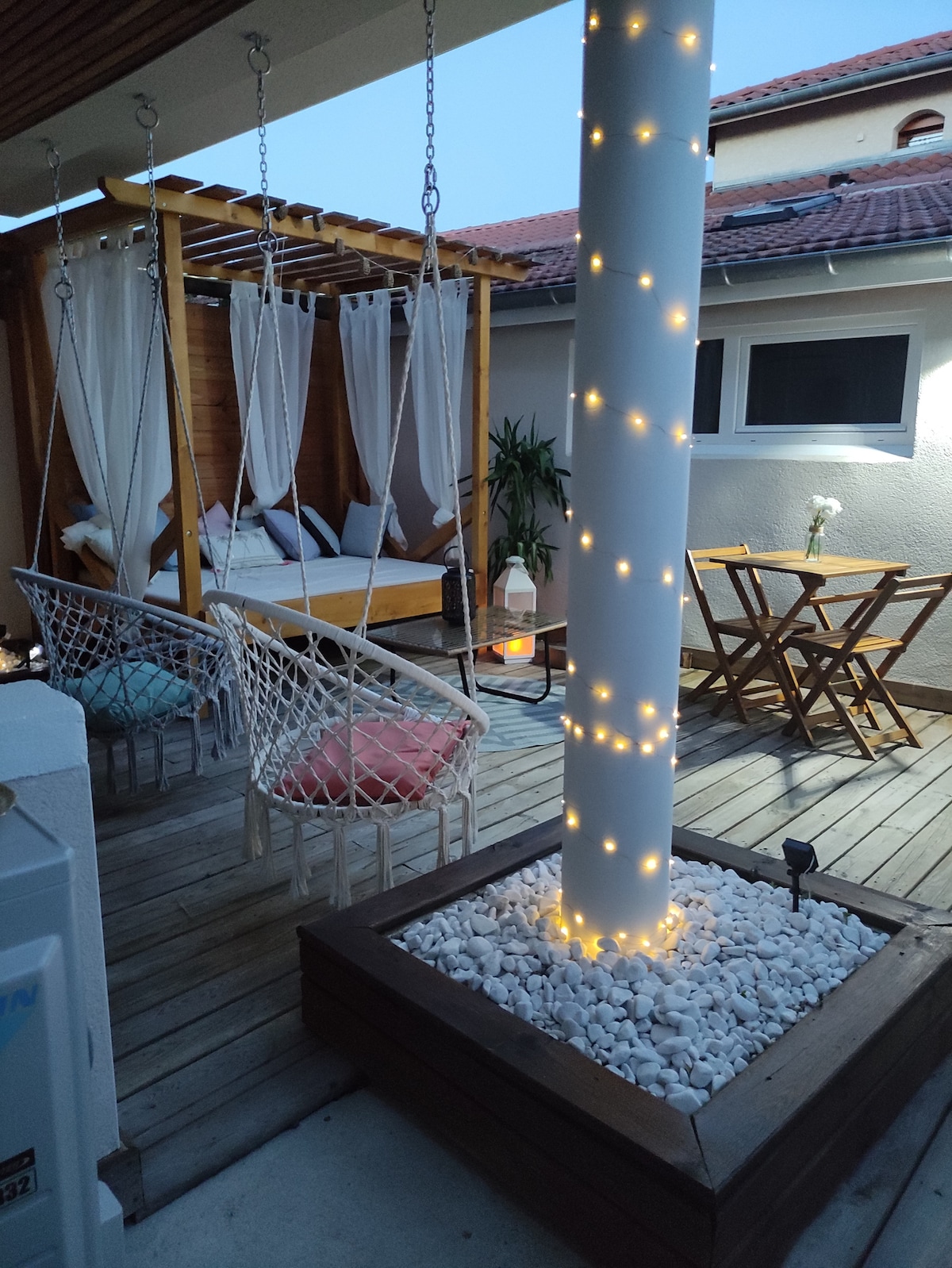 An outdoor space features a wooden structure with a cozy seating area, draped with flowing curtains. Two hanging chairs are positioned beside a small wooden table and chairs. Soft lights are wrapped around a central post, illuminating the area elegantly.