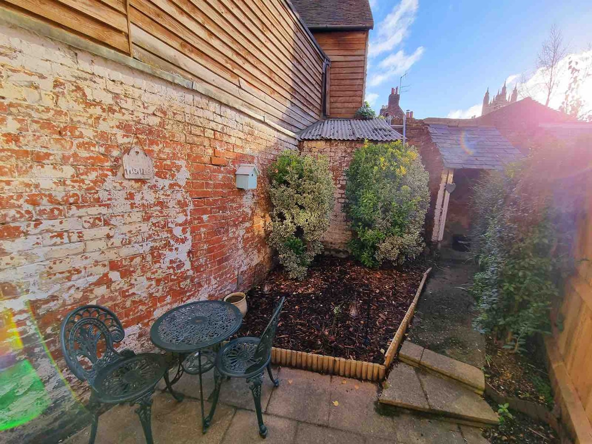 A quaint courtyard is enclosed by textured brick walls, featuring low-maintenance landscaping with shrubs. A small outdoor table with two chairs is positioned on a paved area, offering a peaceful spot for relaxation. Soft sunlight casts gentle shadows across the space.