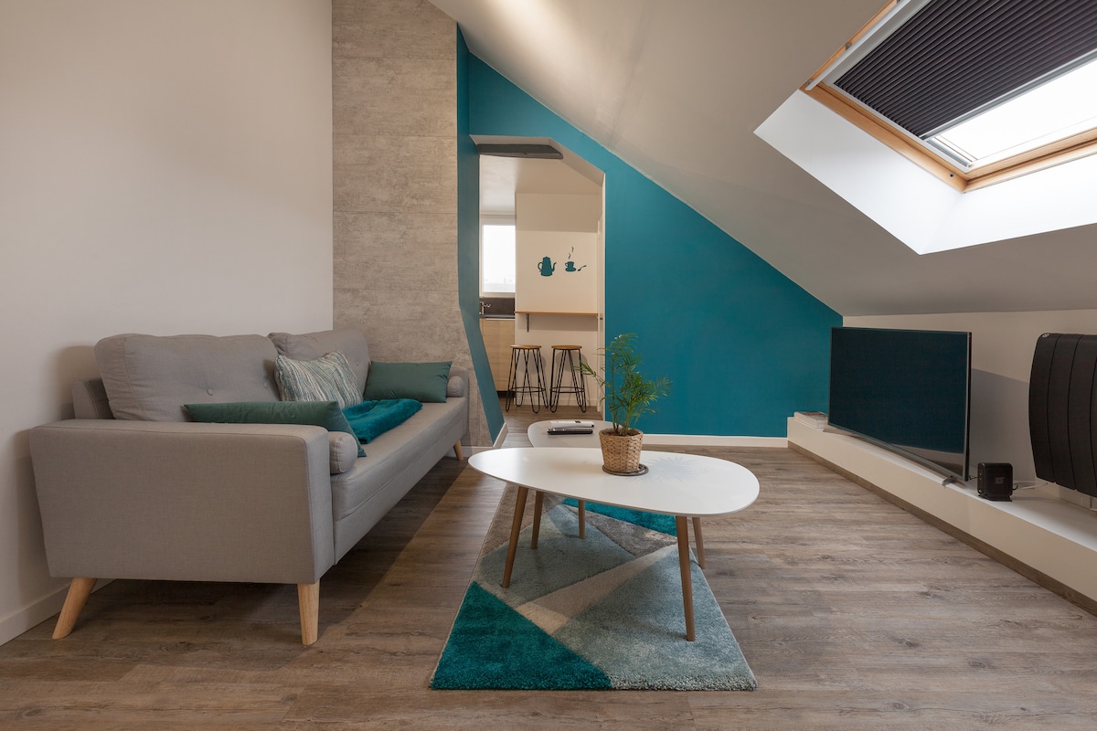 A light-filled living area features a gray sofa adorned with cushions, a white coffee table centered on a patterned rug, and a flat-screen TV positioned against a wall. Angled ceilings and a skylight enhance the space, which is complemented by a vibrant blue accent wall.