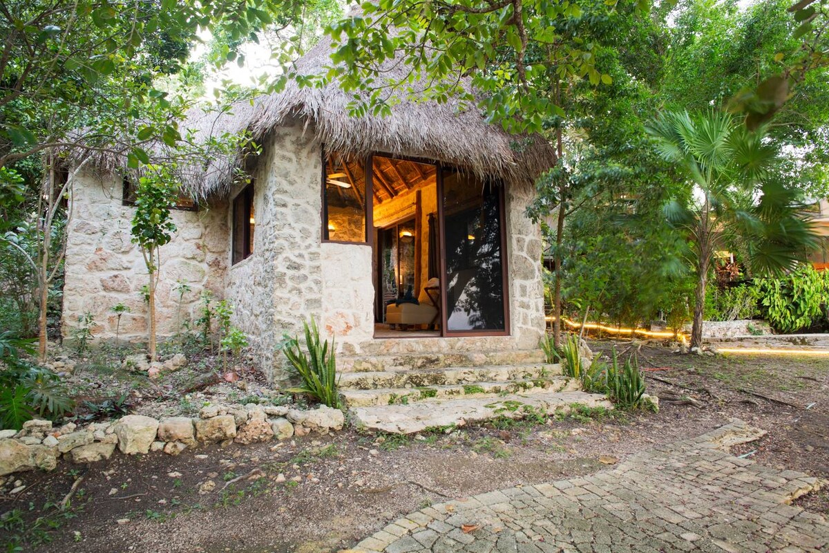 A stone-built bungalow is surrounded by lush greenery, featuring a thatched roof and spacious glass doors. Stone steps lead to the entrance, inviting guests to the serene interior. Soft natural light highlights the bungalow's inviting ambiance, framed by tropical plants and trees.