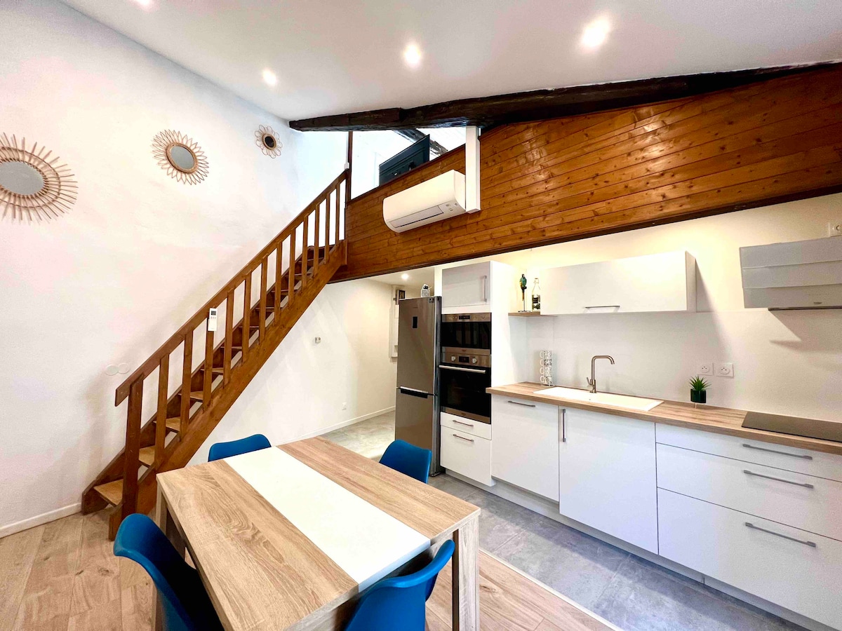 A well-equipped kitchen is showcased alongside a dining area. The space features sleek cabinetry, modern appliances, and a table with chairs. Light from windows enhances the airy feeling, while a wooden staircase adds depth to the room's design.