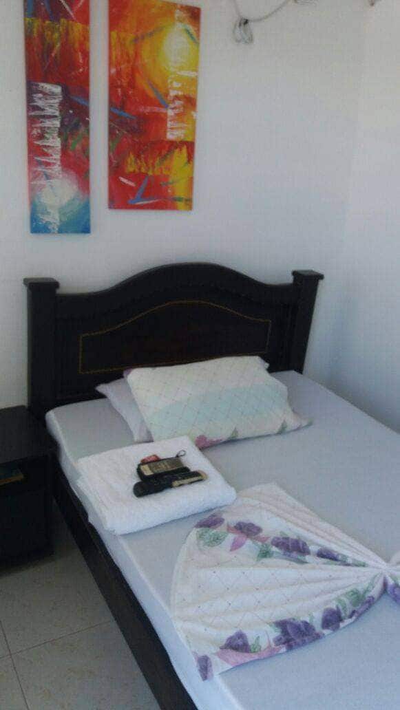 A cozy sleeping area features a dark wooden bed with an unmade mattress, two pillows, and a folded blanket with floral patterns. A bedside table holds a phone. Abstract paintings in vibrant colors are displayed on the wall above the bed.