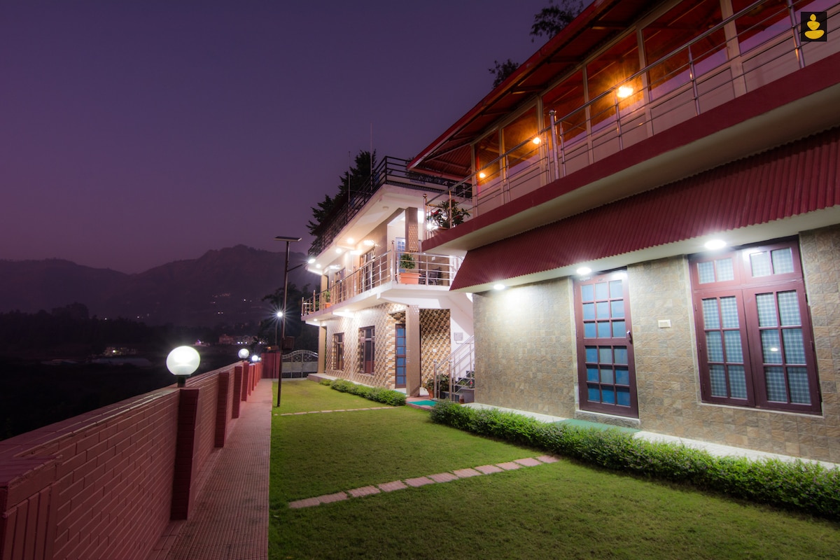 The exterior of the cottage is illuminated in warm tones at dusk, showcasing its multi-level structure and a well-manicured lawn. Lounge areas are accessible, with large windows reflecting the surrounding nature and mountains in the background.