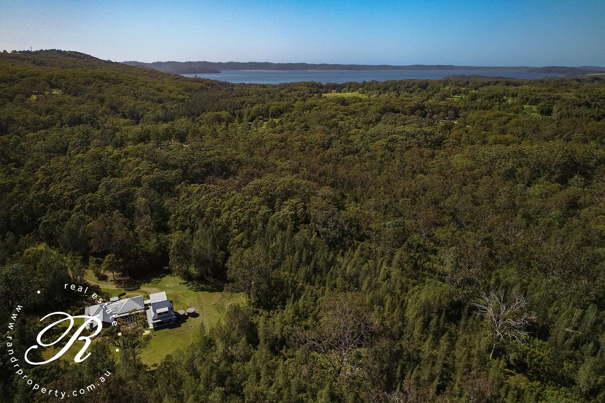 An aerial view captures a secluded house surrounded by dense Australian bushland. The expansive greenery contrasts with the clear blue waters of the nearby lake, offering a serene and private setting. The landscape is dotted with trees, enhancing the sense of tranquility.