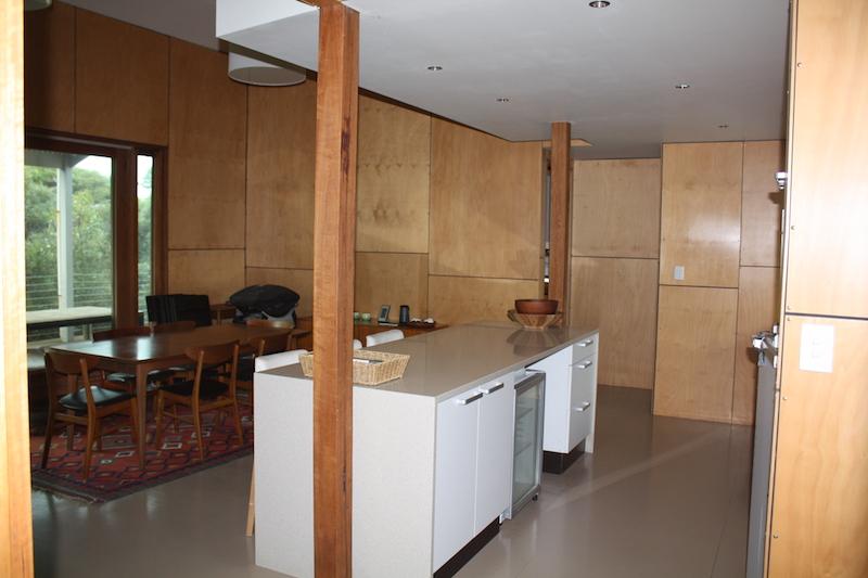 An open-plan kitchen area is highlighted, featuring a central island with a sink and seating. Natural wood paneling decorates the walls, while a dining set with wooden chairs sits nearby. Large windows allow for ample natural light, enhancing the spaciousness of the environment.