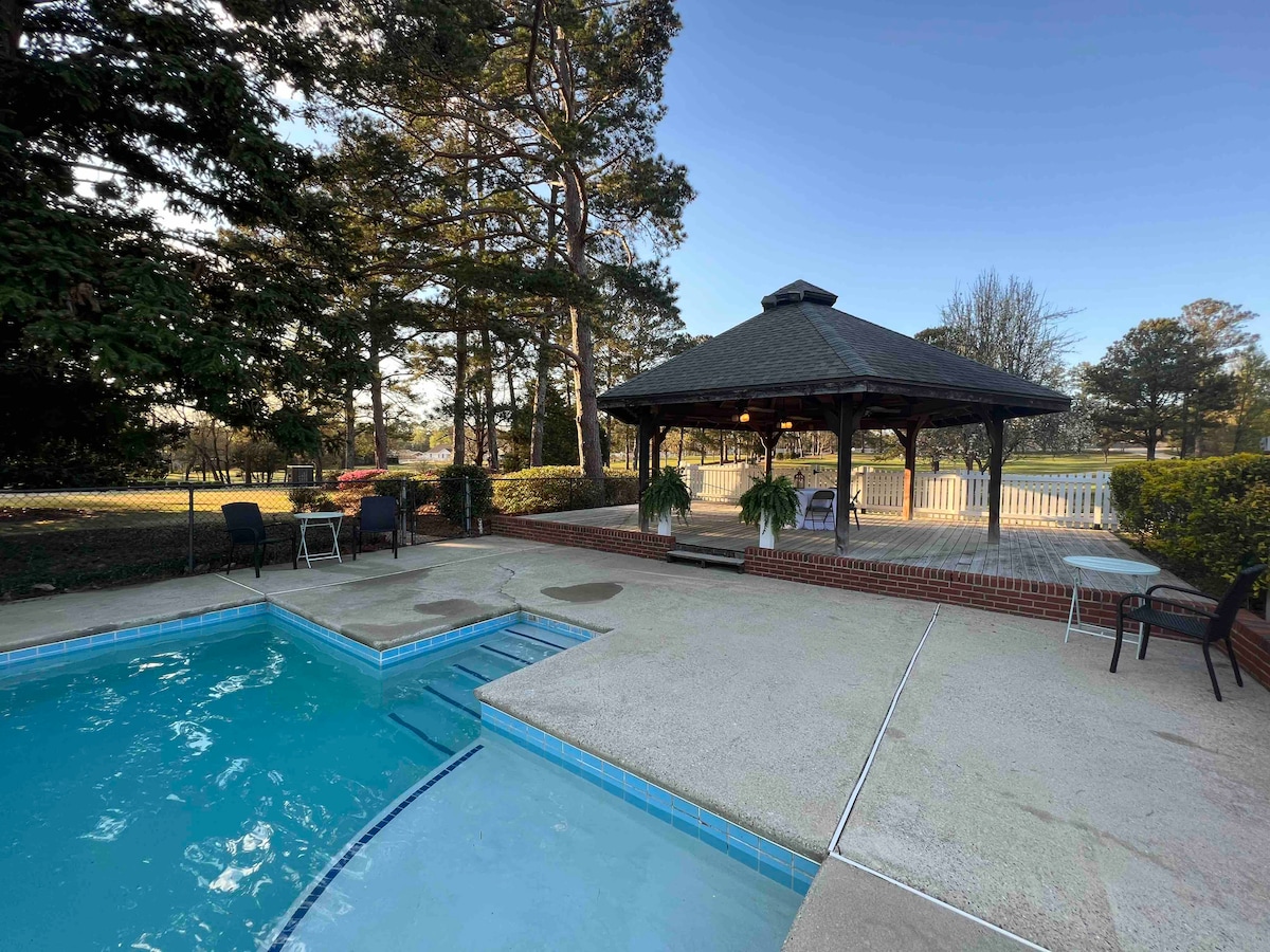 A swimming pool with a clear blue surface is complemented by a nearby gazebo, featuring a wooden structure with seating. Lush greenery and trees surround the area, providing an inviting outdoor setting. The space offers a serene view of the open landscape.