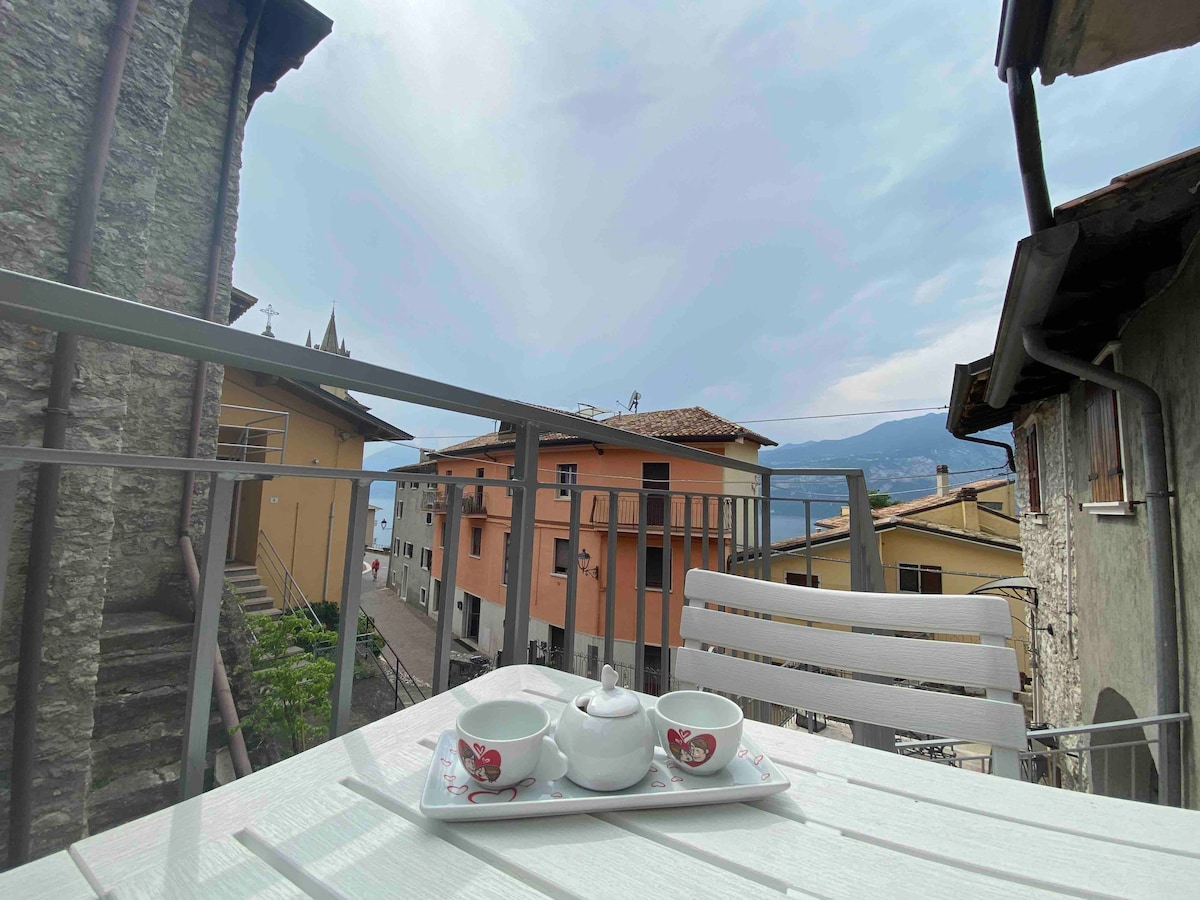A small terrace features a white table set with two matching cups and a sugar bowl, ideal for enjoying a meal or drink. View includes adjacent colorful buildings and a glimpse of the lake in the distance under an overcast sky.