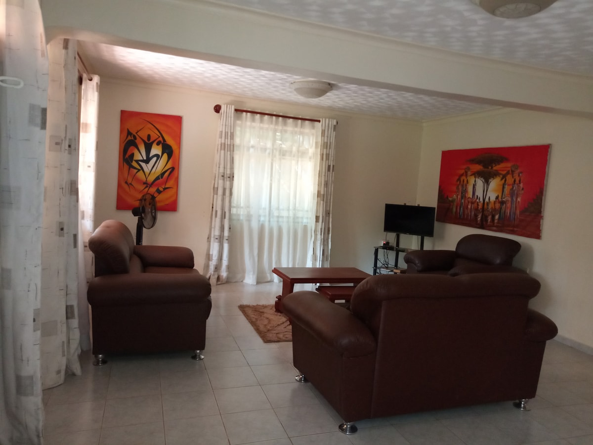 A spacious living area features two brown leather sofas arranged around a wooden coffee table. Bright wall art adds color to the light walls, while sheer curtains frame large windows allowing natural light to fill the space. A small television is positioned in the corner.