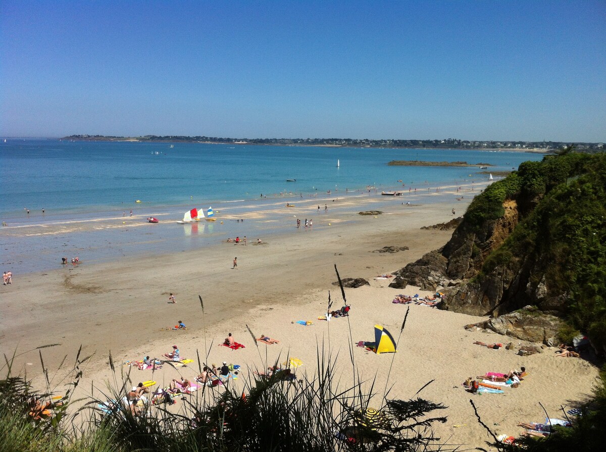 A sandy beach stretches along the shoreline, dotted with colorful umbrellas and beachgoers enjoying the sun. The clear blue water reflects the bright sky, and rocky formations are visible in the distance, adding a natural element to the tranquil scene.