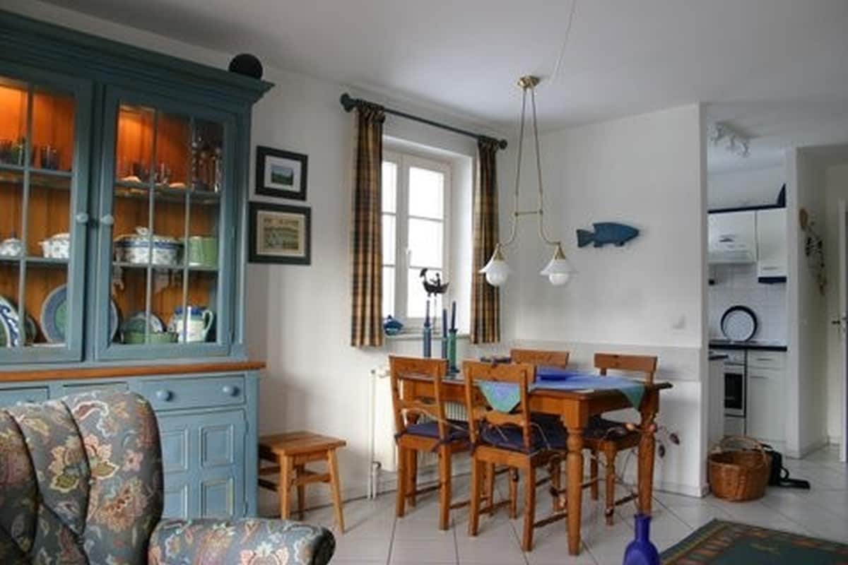 A dining area is presented with a wooden table and four chairs, accented by a blue cabinet displaying ceramic dishware. A window allows natural light to fill the space, while light fixtures hang above the table. A portion of the kitchen is visible in the background.