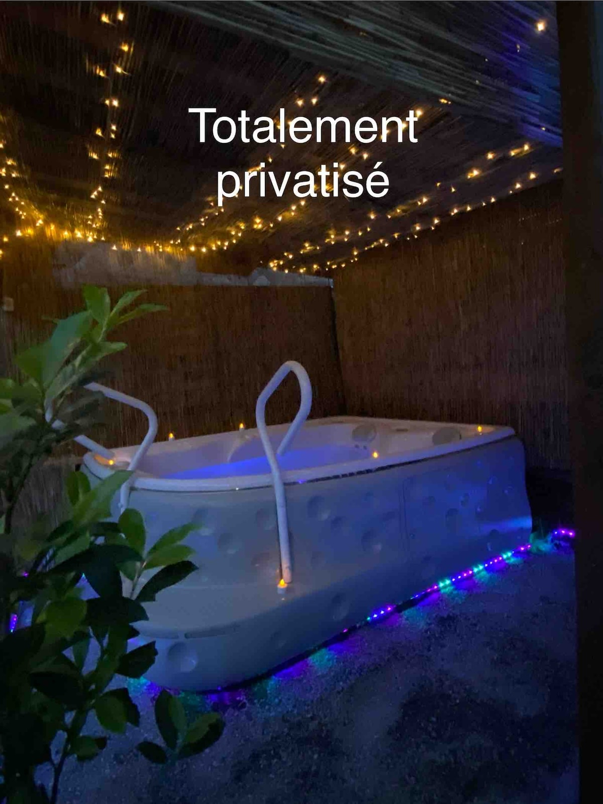 A private jacuzzi is positioned beneath a canopy, surrounded by soft ambient lighting. The tub is illuminated with blue and white lights, creating a serene atmosphere. Lush greenery frames the space, contributing to an intimate and secluded setting.