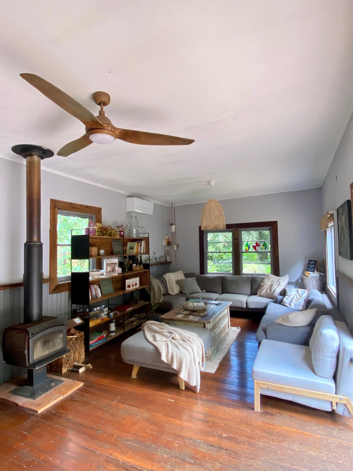 A comfortable living area is featured with multiple seating options, including a gray sectional sofa and separate accent chairs. Natural light filters through the windows, while a fan hangs above. A wooden bookshelf displays various items, and a wood-burning stove adds warmth.