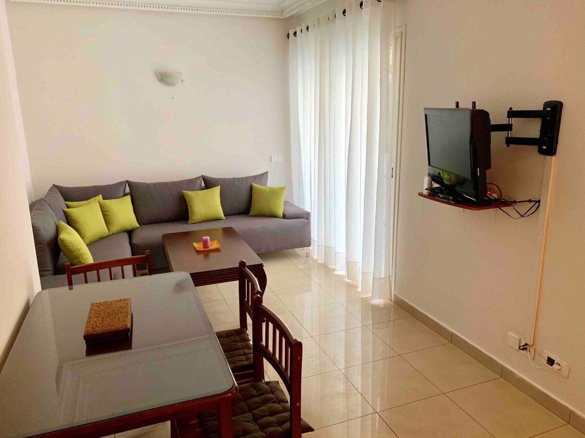 A light-filled living area features a gray sectional sofa with green accent pillows. A wooden dining table is positioned nearby, complemented by four matching chairs. A wall-mounted television is visible, alongside soft white curtains that allow natural light to enter the space.