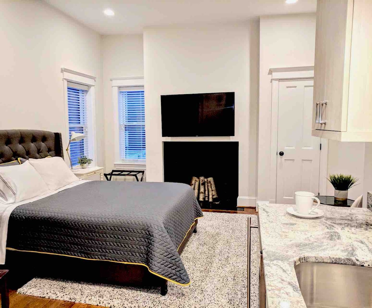 A stylish studio suite features a spacious queen bed covered with a gray quilt and gold trim. A modern flat-screen TV is mounted above a decorative fireplace. Natural light filters through two window panes, enhancing the cozy atmosphere. A kitchenette with granite countertops is visible on the right.