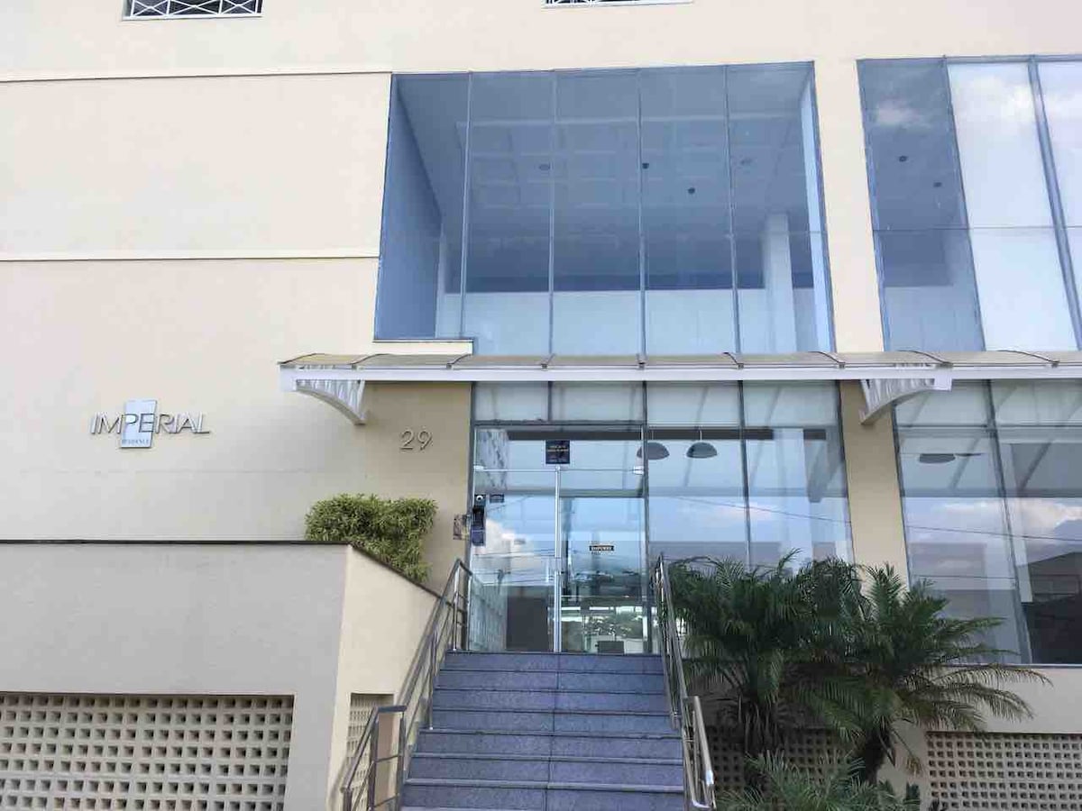 The building entrance is characterized by a modern facade with large glass doors and a prominent signage displaying 'IMPERIAL.' A set of stairs leads up to the entrance, flanked by minimalist landscaping with greenery.