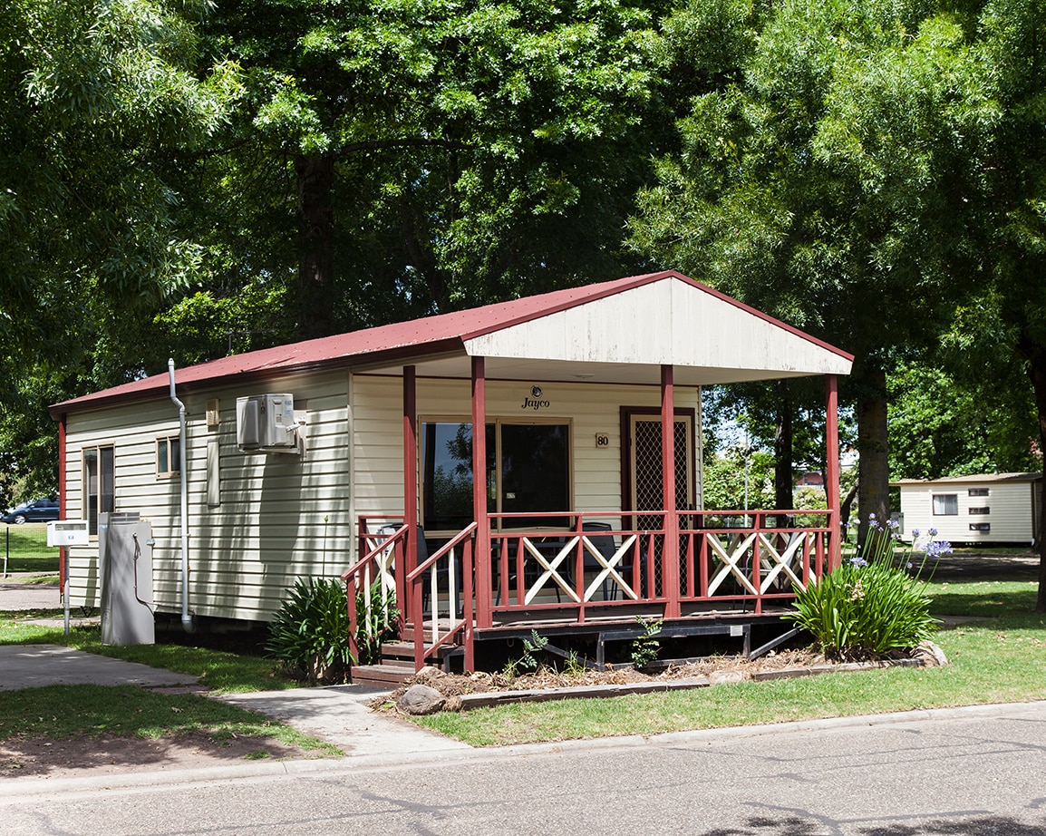 A wooden cabin with a covered porch is framed by tall trees and landscaped greenery. The cabin features large windows and a welcoming entryway with wooden railings, providing a glimpse into a comfortable atmosphere set within a holiday park.