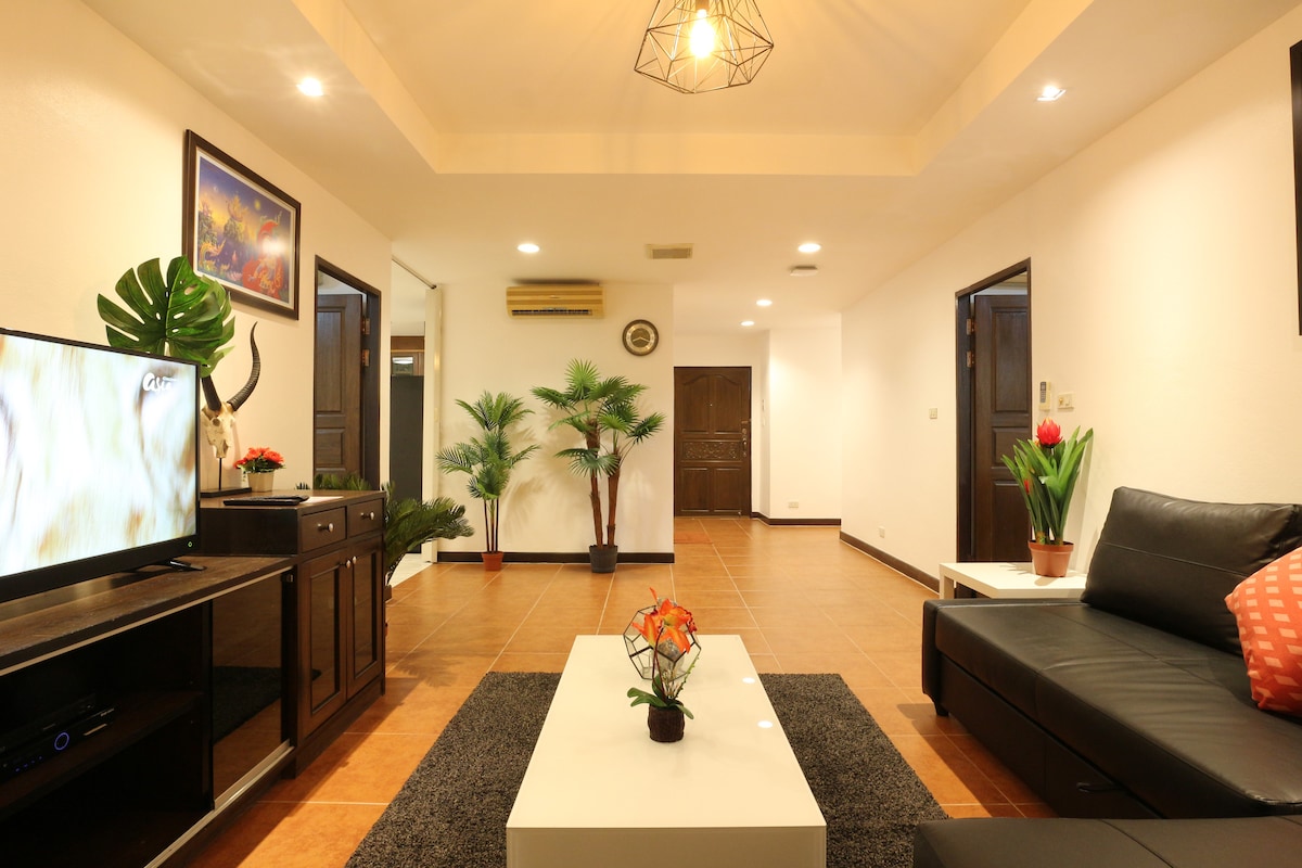 A spacious living area features a dark sectional sofa adorned with decorative pillows. A modern coffee table sits at the center, accompanied by a flat-screen TV mounted on a stylish console. Potted plants add greenery, enhancing the room's inviting ambiance.