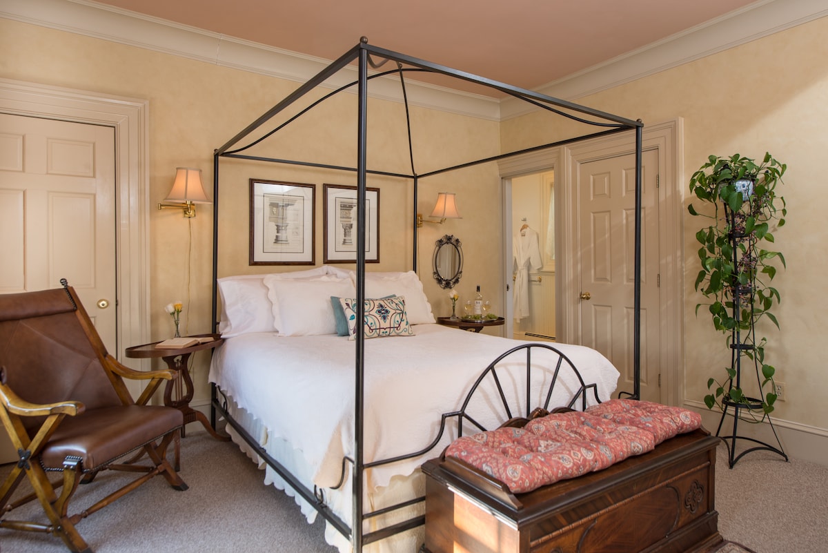 A romantic room is highlighted by a queen canopy bed draped with soft linens. A wooden trunk sits at the foot of the bed, and a plant is positioned in the corner. The space is complemented by framed artwork and bedside lamps, creating an inviting ambiance.