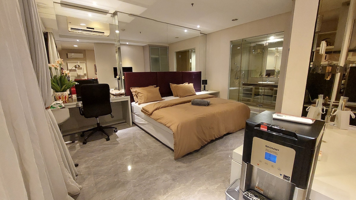 A spacious bedroom area is furnished with a large bed featuring a neutral-toned duvet. A desk with a chair is positioned next to a mirror, while modern fixtures enhance the clean aesthetic. The floor is polished, and a coffee maker is visible on a nearby countertop.