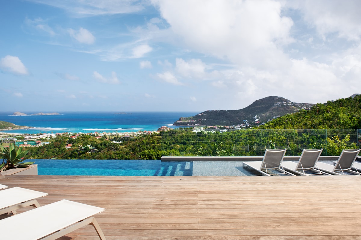 An outdoor terrace features a spacious infinity pool overlooking the expansive Bay of St. Jean. Nearby, several lounge chairs are positioned on the wooden deck, providing a serene view of the lush landscape and vibrant ocean waters.