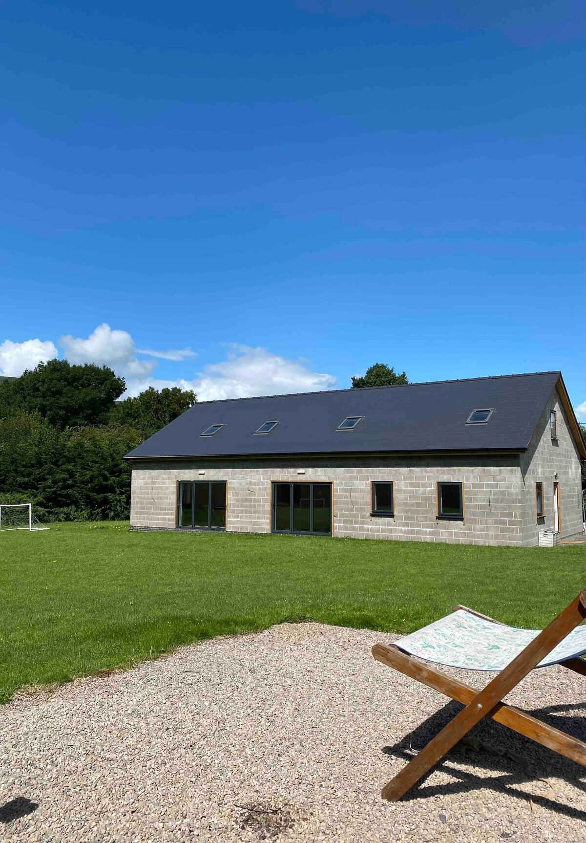 A spacious structure features large glass doors and skylights, set against a backdrop of lush greenery. An open field is visible in front, and a lounger is placed on a gravel area for relaxation. The clear blue sky complements the tranquil setting.
