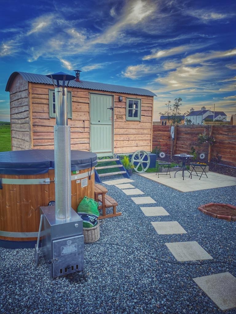 A charming shepherd's hut is set against a vibrant sky, with a pathway leading to a private outdoor area featuring a log-burning hot tub and a bistro table with chairs. The space is surrounded by decorative pebbles and green foliage, enhancing the tranquil atmosphere.
