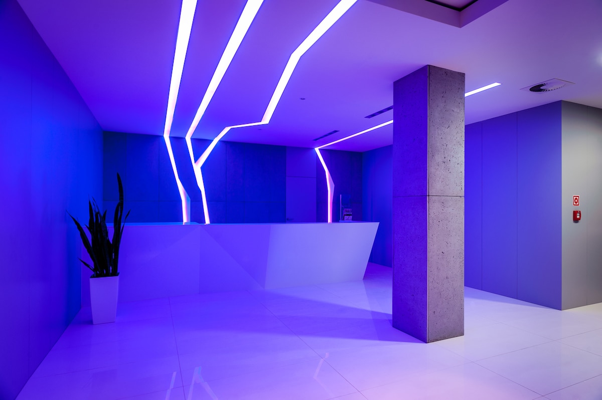 A modern reception area is illuminated by vibrant blue and purple lighting, creating a dynamic atmosphere. A sleek white reception desk is complemented by concrete walls, while a tall plant adds a touch of greenery to the space.