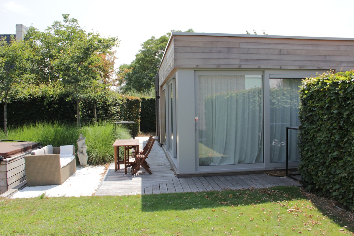 A modern vacation home is surrounded by lush greenery, featuring large glass sliding doors that invite natural light. An outdoor seating area is visible, complete with comfortable chairs and a small dining table, set on a wooden deck adjacent to a landscaped garden.
