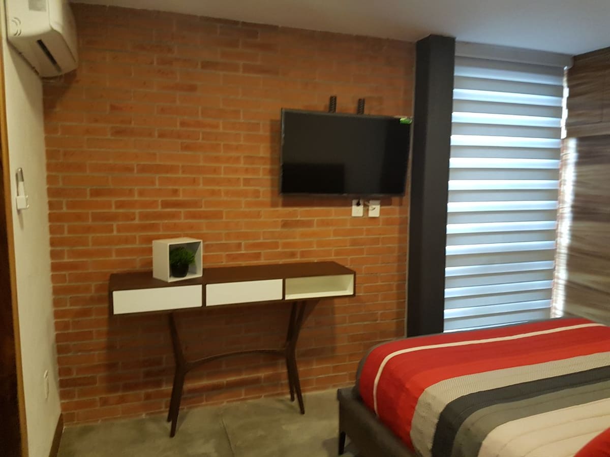 The room features a textured brick wall and a sleek media console with two drawers. A flat-screen television is mounted above. Light filters through layered window coverings, while a bed with colorful striped bedding adds a touch of comfort to the space.