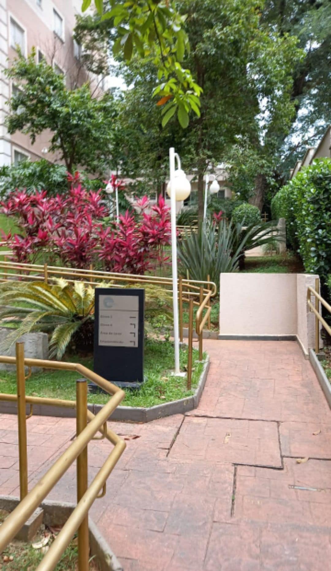 A landscaped pathway meanders through vibrant greenery, featuring lush pink and green plants alongside a railing. A sign is positioned near the walkway, indicating directions. Warm lighting fixtures subtly enhance the inviting atmosphere of the area.