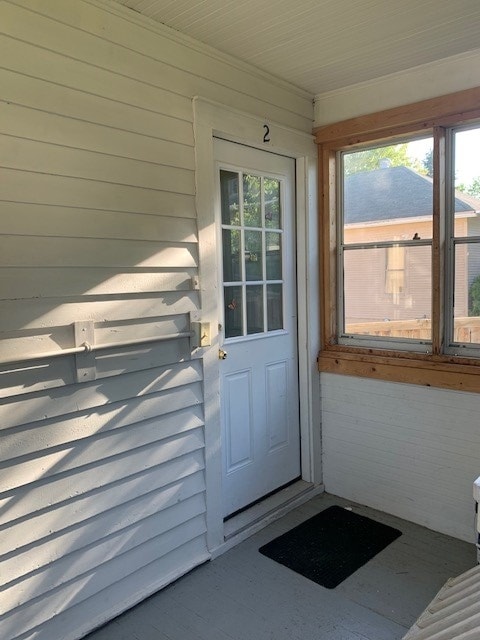 A welcoming entryway is presented with a white door featuring multiple glass panes, allowing natural light to enter. The walls are lined with horizontal white panels, complemented by a gray mat on the floor. A wooden frame enhances the surrounding area of the door.