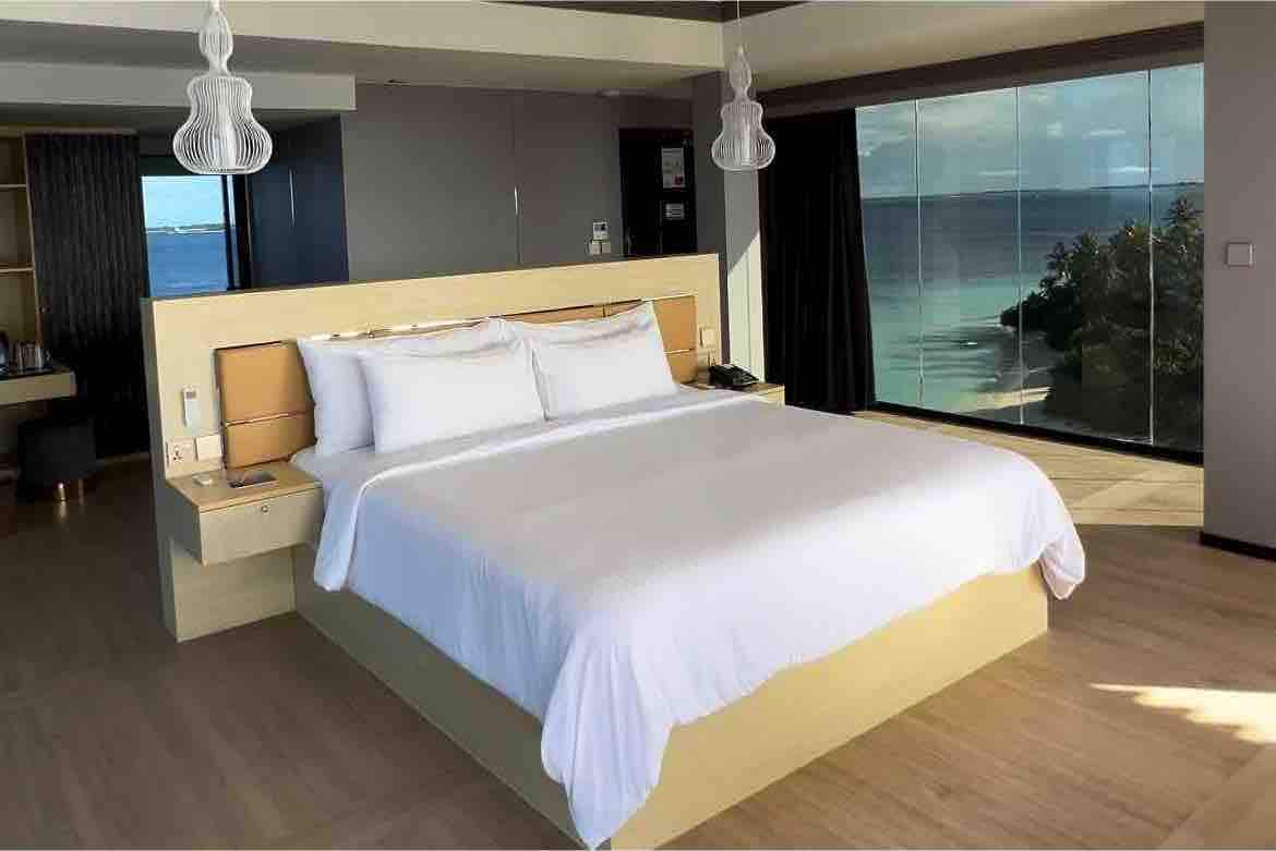 A spacious bedroom features a large bed with crisp white linens, positioned against a backdrop of expansive windows that offer views of the ocean. Soft lighting is provided by modern pendant fixtures, and subtle decor enhances the calming atmosphere.