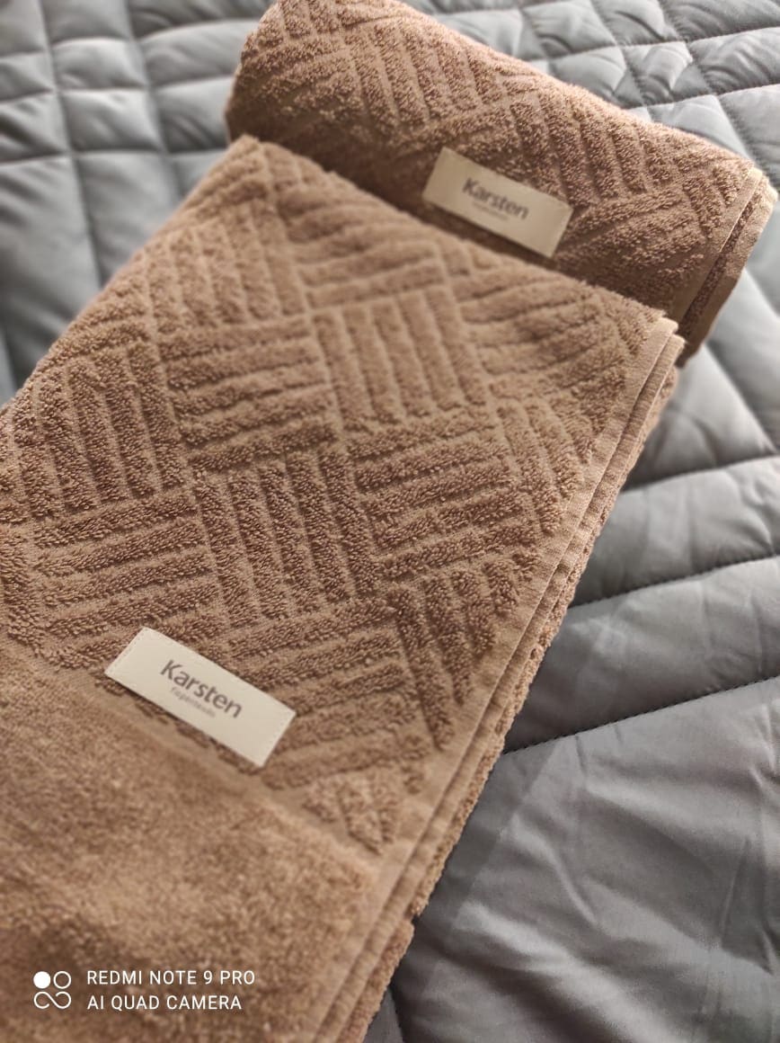 A neatly arranged set of brown towels is displayed atop a grey quilted surface. The towels feature a textured pattern and a small label indicating the brand. The soft fabric and neutral colors create a calm aesthetic.