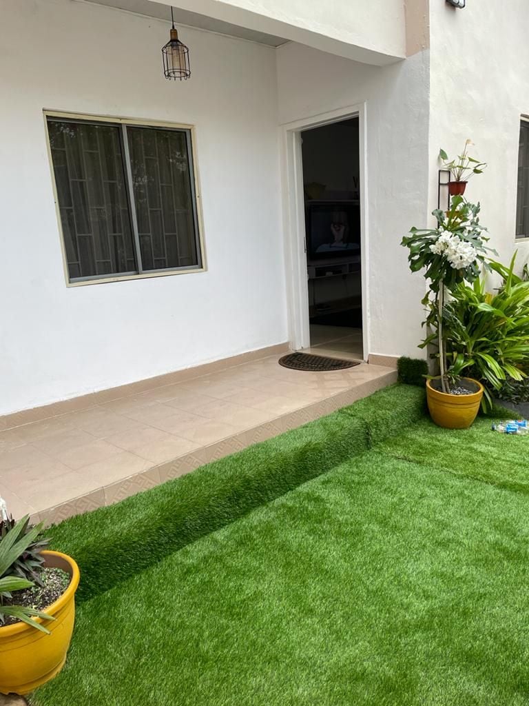 A well-maintained exterior features a small patio area, leading to an entrance door. Lush green artificial grass covers the ground, complemented by potted plants. A window provides natural light, while a warm ambiance is suggested by a hanging light fixture.