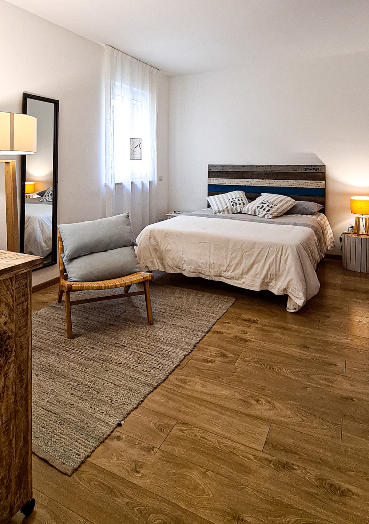 A comfortable bedroom is shown, featuring a spacious bed with simple bedding. Soft lighting is provided by two bedside lamps. A woven area rug lies on the wooden floor, and a chair is positioned near a full-length mirror, creating a cozy atmosphere.
