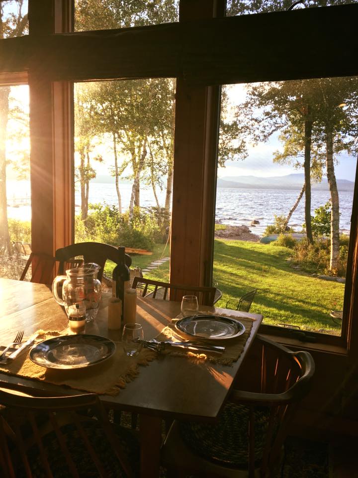 A dining table is set with plates and glassware, positioned near large windows. Warm sunlight filters through the trees, illuminating the outdoor lake view, showcasing a blend of lush greenery and shimmering water.