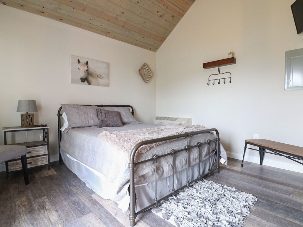 A cozy sleeping area is furnished with a queen bed, draped in soft linens, and placed against a light-colored wall. A rustic horse-themed artwork complements the decor. Natural light enters through the window, illuminating the wooden ceiling and textured rug positioned beside the bed.