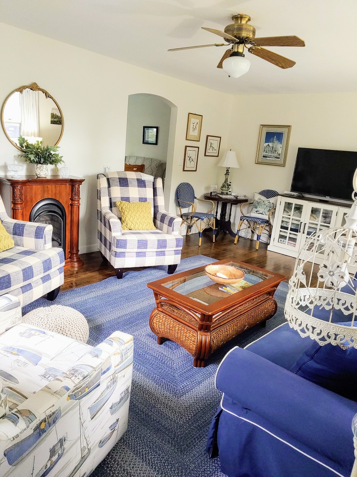 A cozy living area showcases a blend of comfortable seating options, including two checked armchairs and a blue sofa. A decorative coffee table sits at the center, accompanied by a chandelier. Light oak flooring complements the space, with art adorning the walls and a television present.