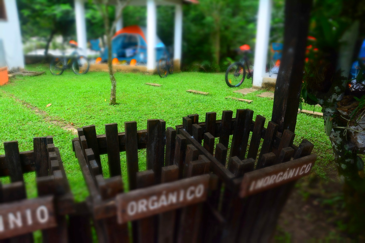 Two wooden waste bins are labeled for organic and inorganic waste, positioned on a grassy area. In the background, several tents are visible under a covered space among lush greenery, contributing to a natural environment.