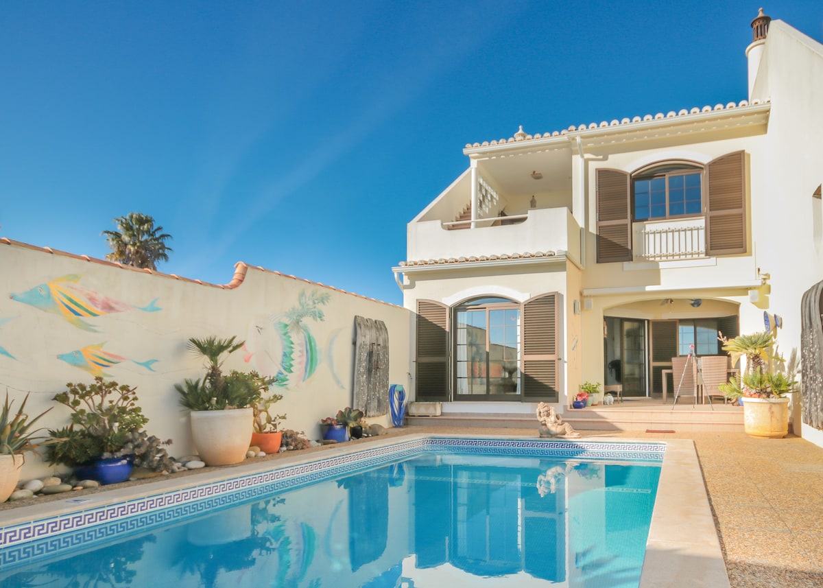 A well-maintained private outdoor area features a saltwater pool surrounded by various potted plants. The house includes a charming mural on one wall and ample space for relaxation. Large windows hint at comfortable indoor living spaces, with sun-kissed water reflecting the clear blue sky.