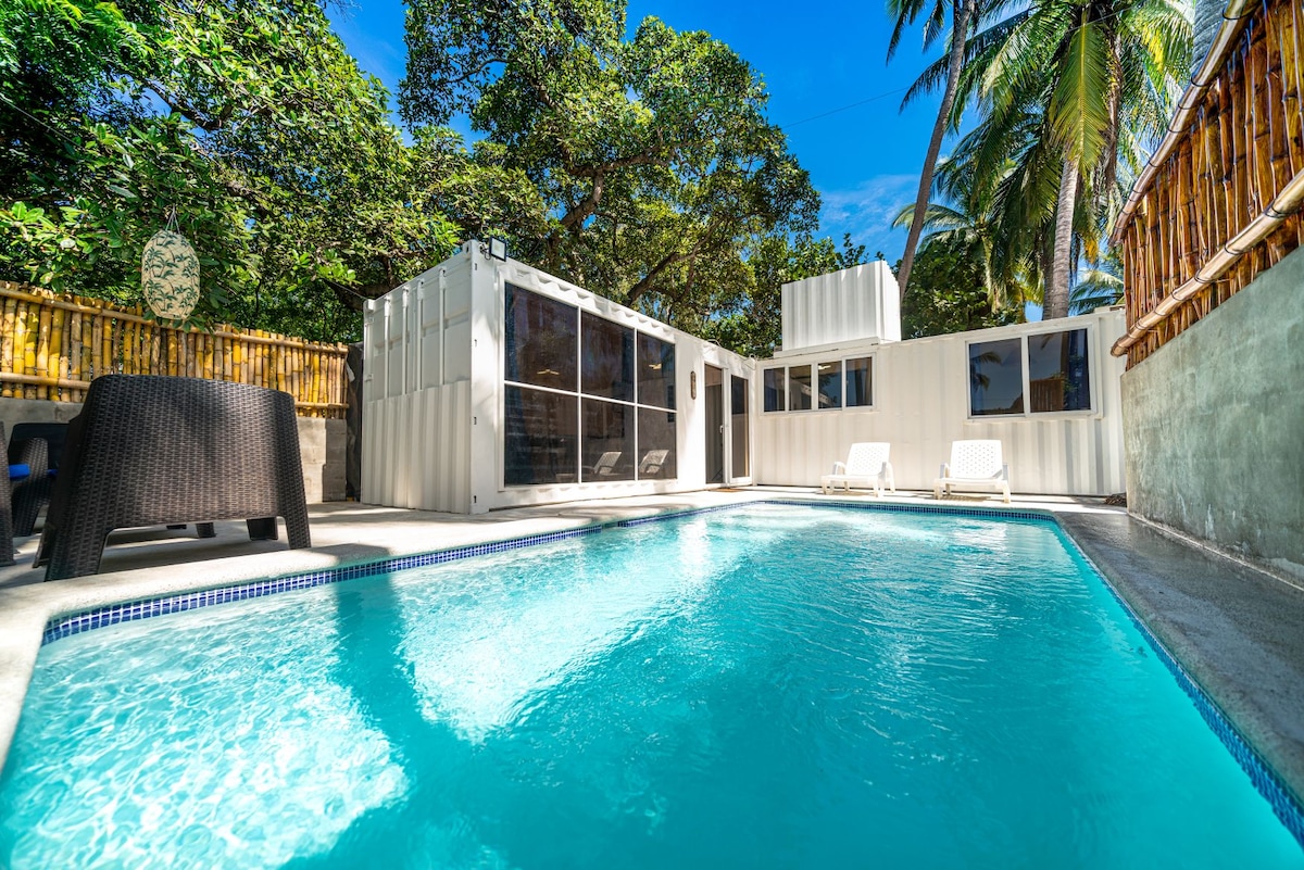 A refreshing pool reflects the sky, bordered by lounge chairs and landscaped greenery. The surrounding area features a modern building with large glass panels, providing an inviting atmosphere. Lush palm trees frame the scene, enhancing the sense of relaxation in the tropical setting.