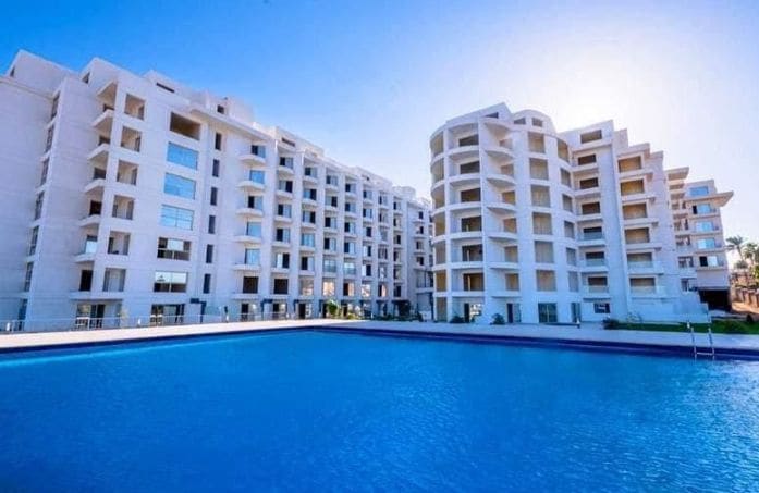 A modern building complex is situated next to a clear blue pool. The architecture features multiple levels with balconies and large windows, allowing natural light to fill the space. Lush greenery is visible around the pool area, enhancing the tranquil setting.