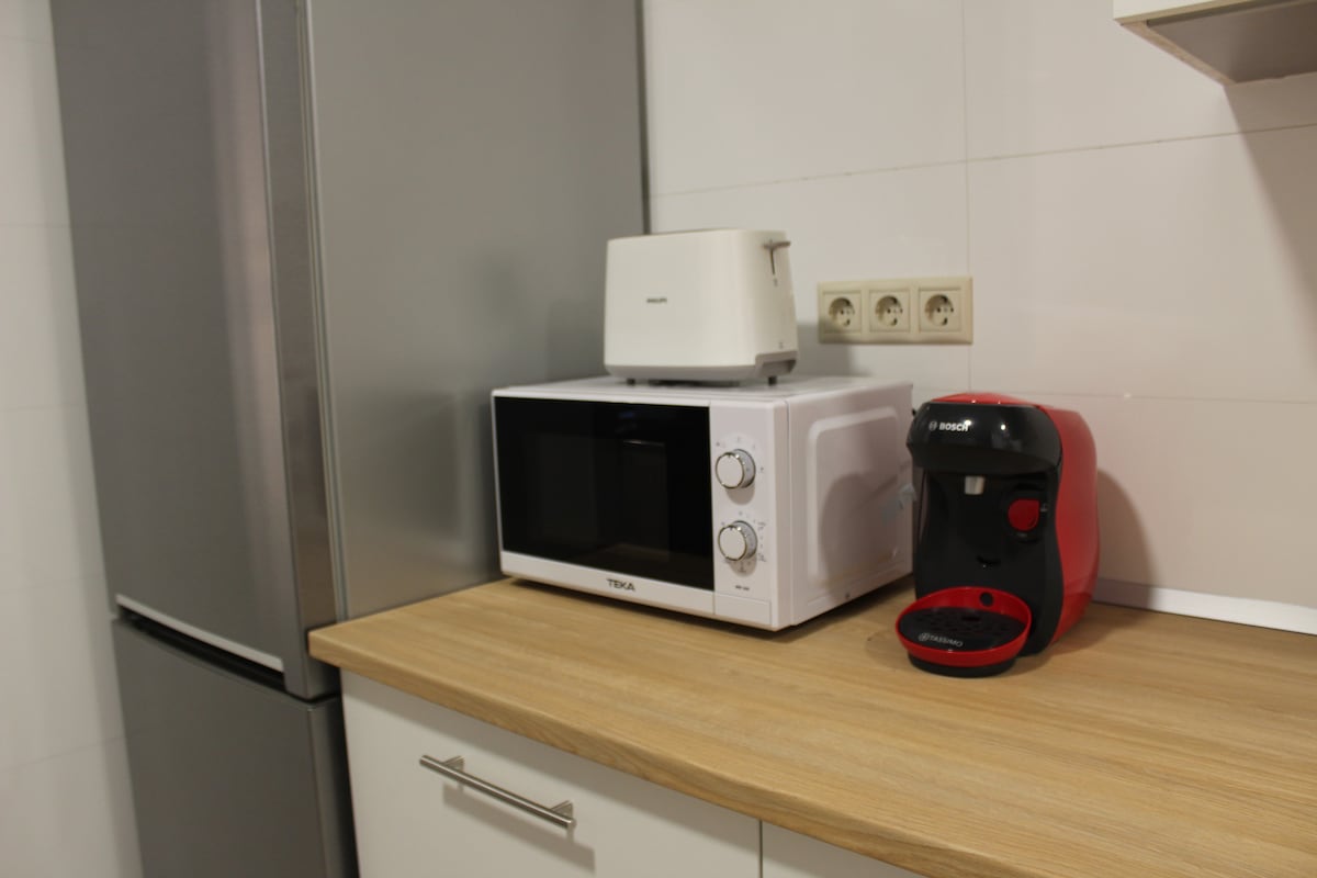 A modern kitchen countertop is featured with essential appliances, including a white microwave, a sleek silver toaster, and a red coffee maker. The wooden surface complements the minimalist design, while the surrounding cabinetry offers additional storage options.