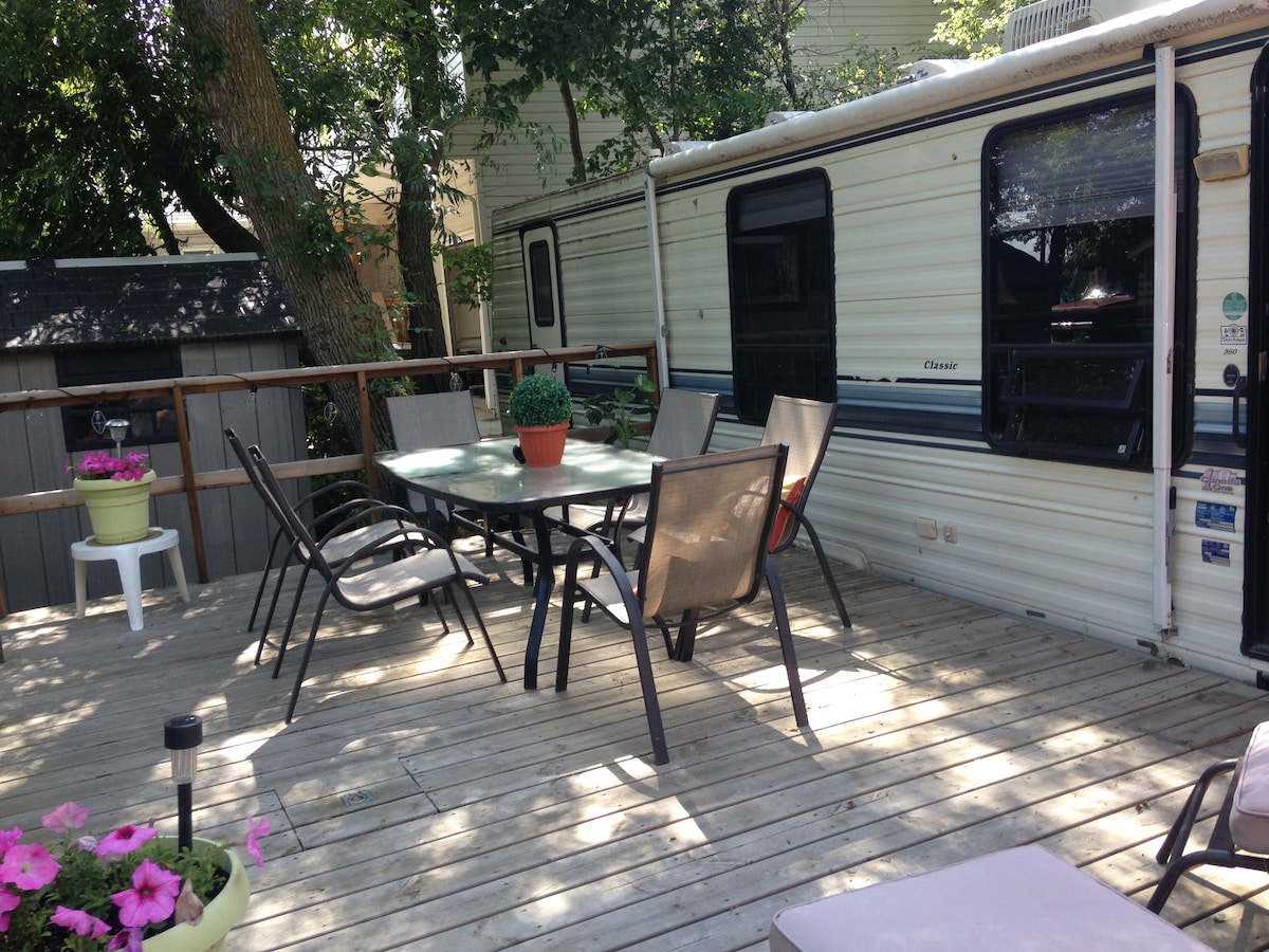 A spacious deck is featured around the trailer, equipped with a dining table surrounded by five chairs. Potted plants are placed on the table and nearby, adding greenery to the outdoor space. Sunlight filters through the trees, creating a serene atmosphere.