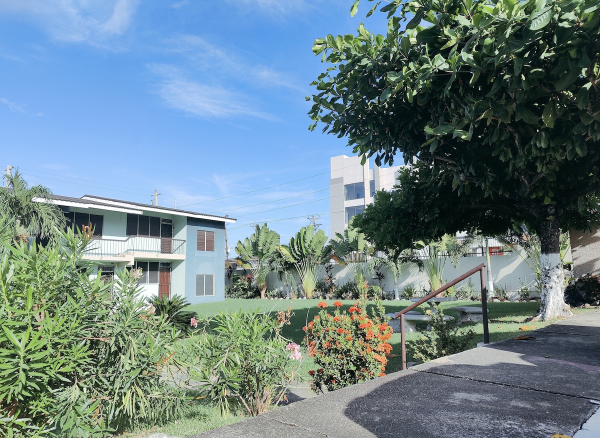 The image displays a green courtyard surrounded by vibrant foliage and blooming flowers. Two multi-story structures are visible, showcasing a blend of modern and traditional architecture. A sturdy staircase leads down from the foreground, providing access to the grassy area.