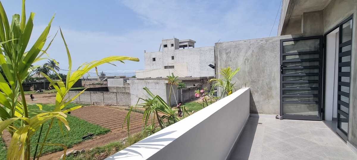 A spacious balcony with modern railings features potted plants and a clear view of the surrounding landscape, including fields and distant buildings. The ocean can be glimpsed under a bright blue sky, providing a serene backdrop.