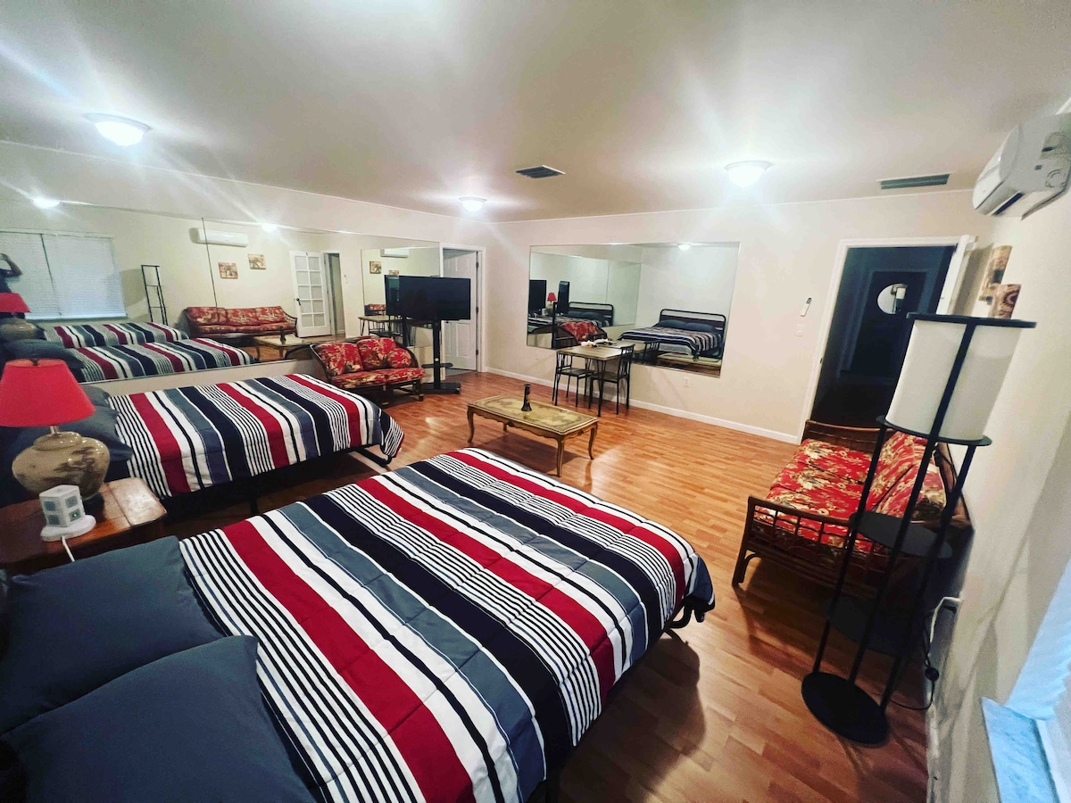 A bright, spacious room is equipped with two striped beds, complemented by a mirror and a small table. A television is positioned nearby, and wooden flooring adds warmth to the space. Additional seating is provided with a wicker chair and a lamp stands at the side.