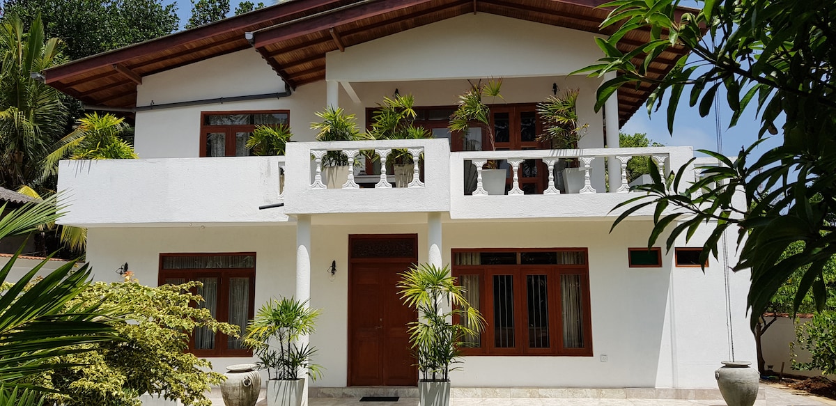 The exterior of a spacious villa is showcased, featuring a welcoming entrance with a wooden door framed by large windows. Lush greenery surrounds the building, and potted plants are placed along the balcony railing, creating an inviting atmosphere.